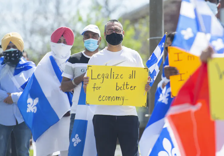 A group of people wearing face masks hold up Québec flags and a protest sign that says 'Legalize all for better economy'