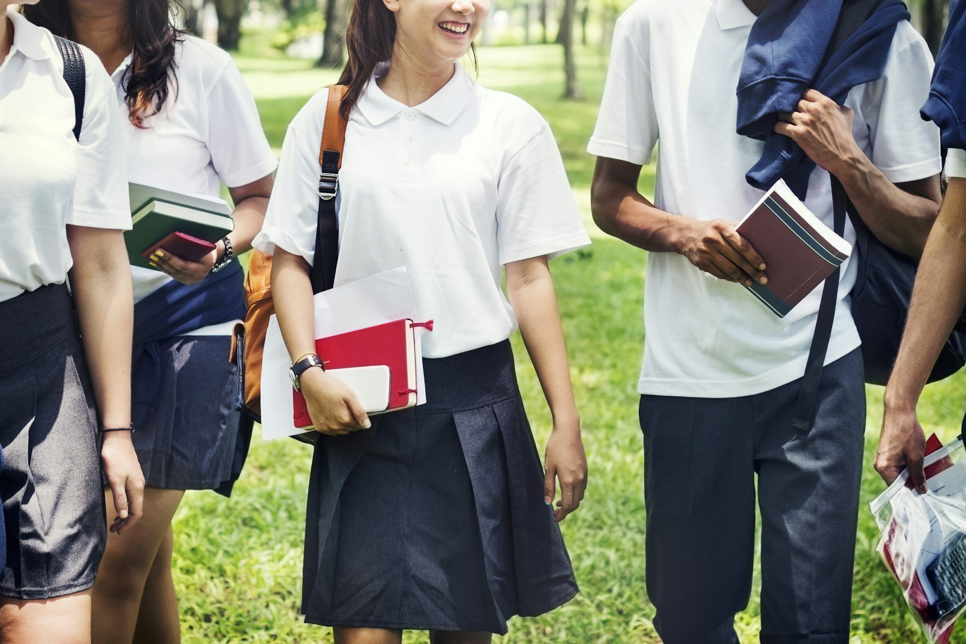 A group of teenagers in navy skirts and pants and white shirts walk, carrying books across grass.