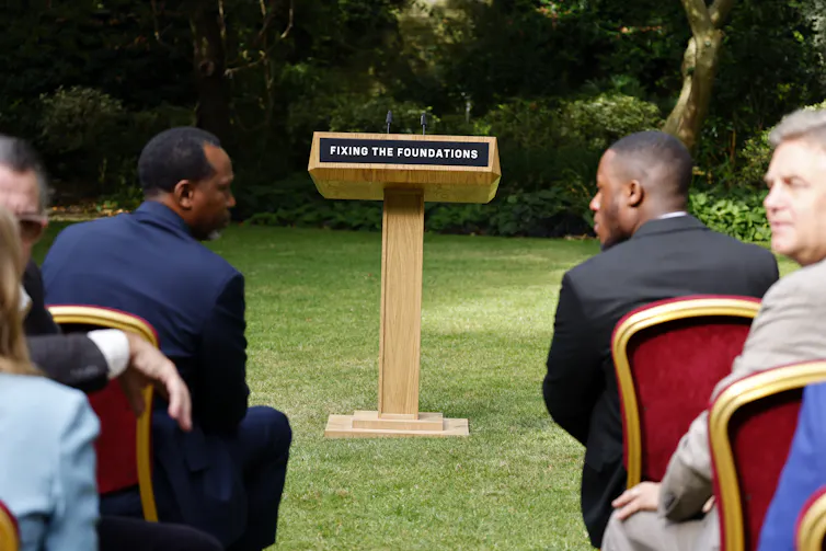 Two men in an audience talking to each other across an aisle with a podium in front of them.