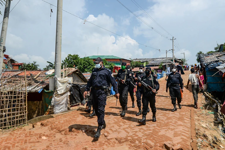 A group of armed men in black uniform walk down a dustry track with shacks in the back.