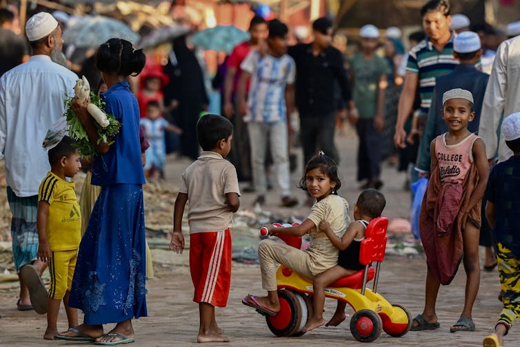 A girl on a tricycle is surrounded by other children in a refugee camp.
