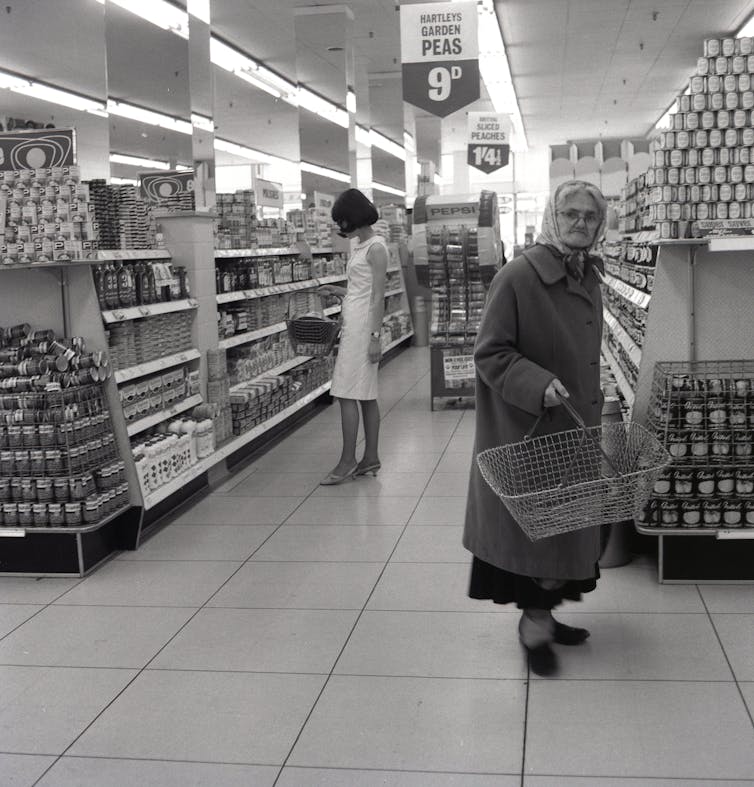 An old lady with shopping basket inspects the new 'self-service' shopping shelves, with a younger woman behind her.