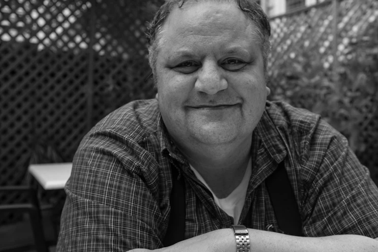 A smiling man wearing braces over a checked short-sleeved shirt, arms folded