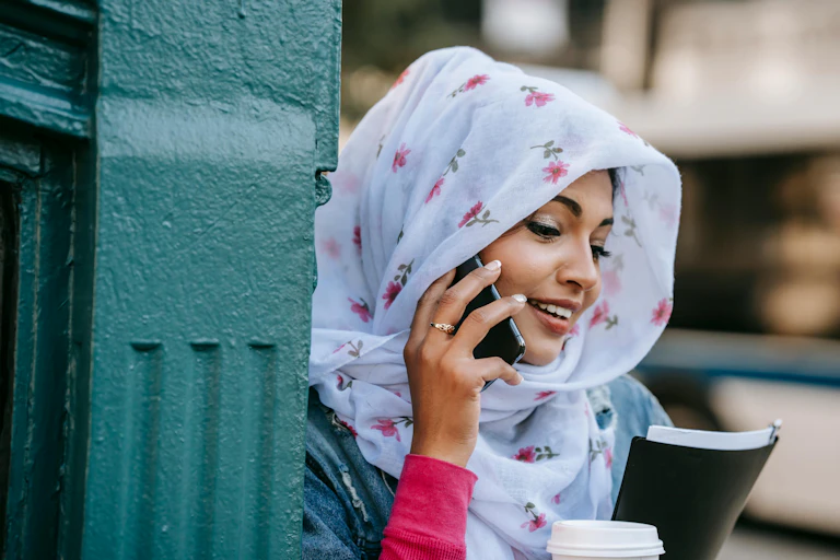 A young woman in a white floral head scarf smiling talking on a phone.