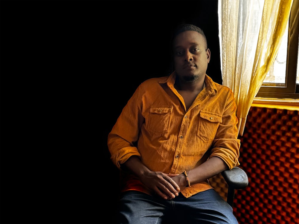 A man sitting on a chair, surrounded by shadow and looking down the lens.