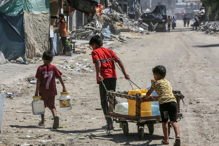 Three Palestinian children collect drinking water in a war-damaged street.
