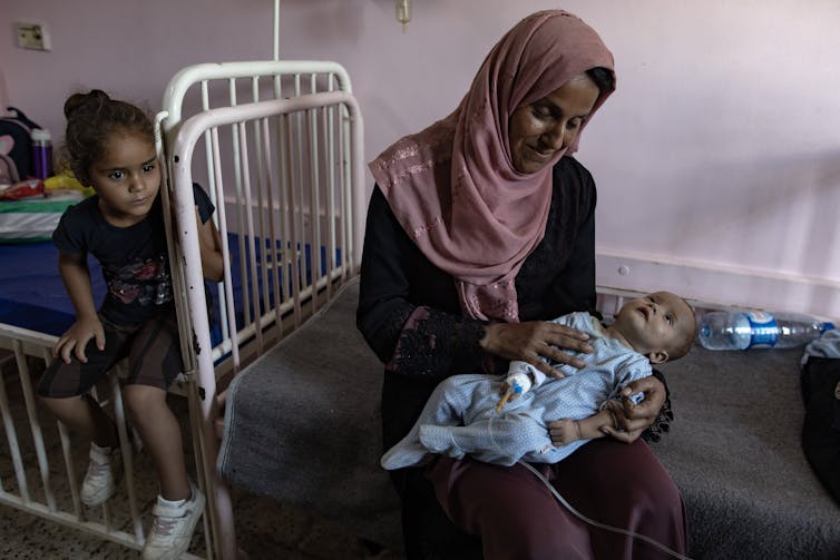 A mother sat on the floor with a baby in her lap as another child sits on a bed next to them.