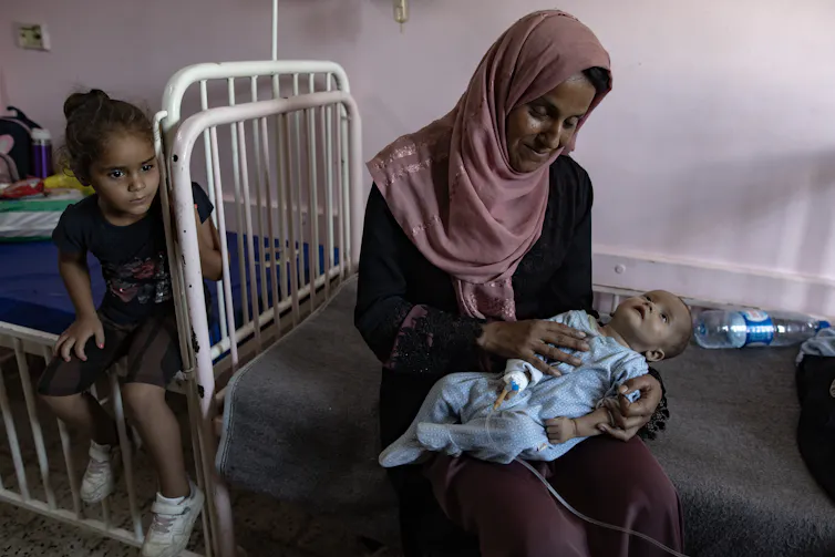 A mother sat on the floor with a baby in her lap as another child sits on a bed next to them.
