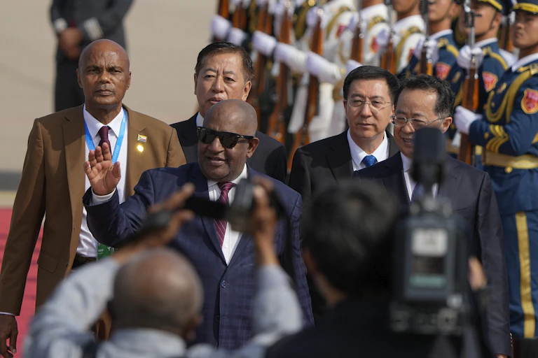 A man wearing sunglasses waves as he walks past a military guard.