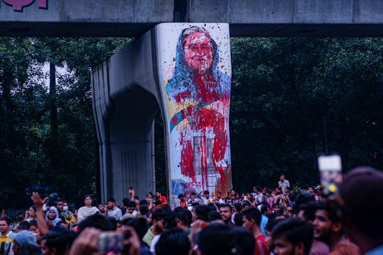 Protesters gather in front of a picture of Sheikh Hasina that has been covered in red paint.