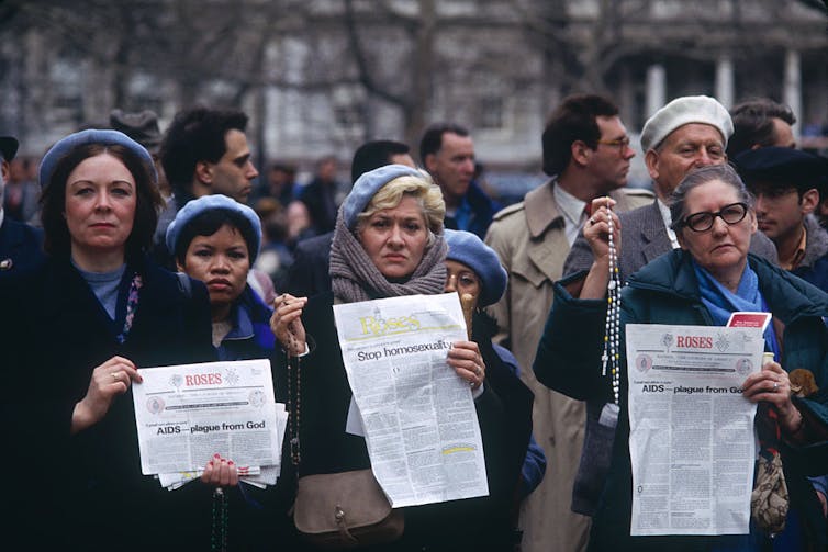 Four women stand at the front of a crowd, holding white leaflets; one of them also holds a white rosary.