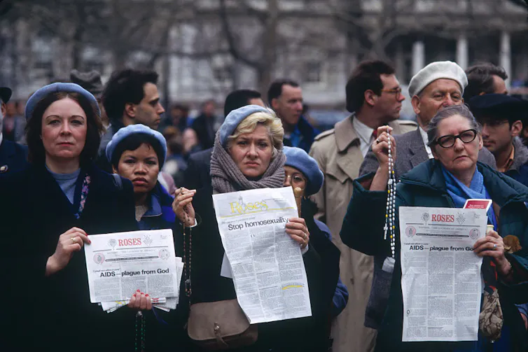 Four women stand at the front of a crowd, holding white leaflets; one of them also holds a white rosary.