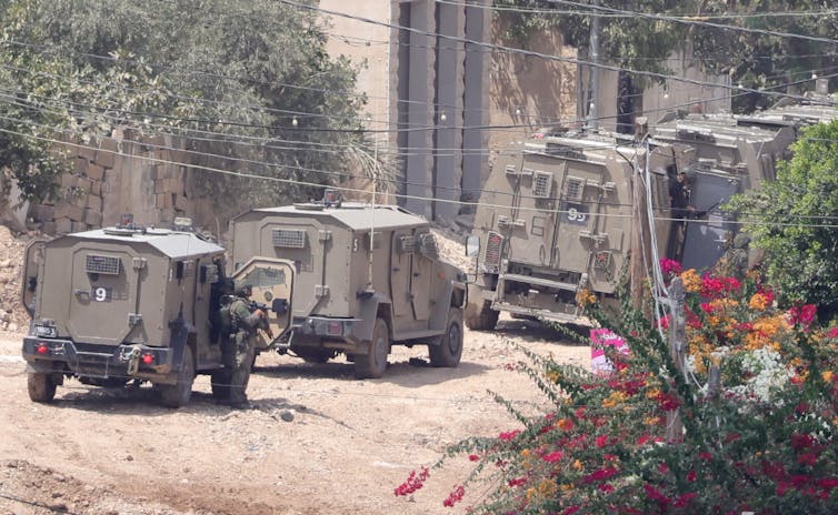 A column of Israeli armoured vehicles on a dirt track in a city.