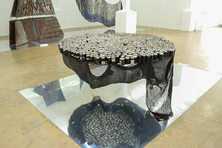 An art gallery with a round sculpture suspended from the roof bearing multiple homemade lanterns, in turn reflected by mirrors beneath it on the gallery floor.
