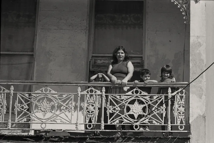 A woman and her children look down from the balcony of an old terrace house.