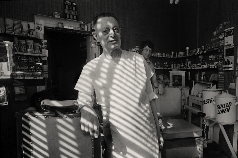 A barber poses in a white jacket in his crammed shop.