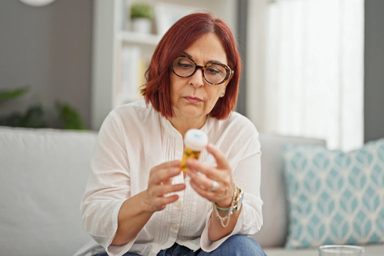 A middle-aged woman examining a jar of tablets while sitting on a couch..
