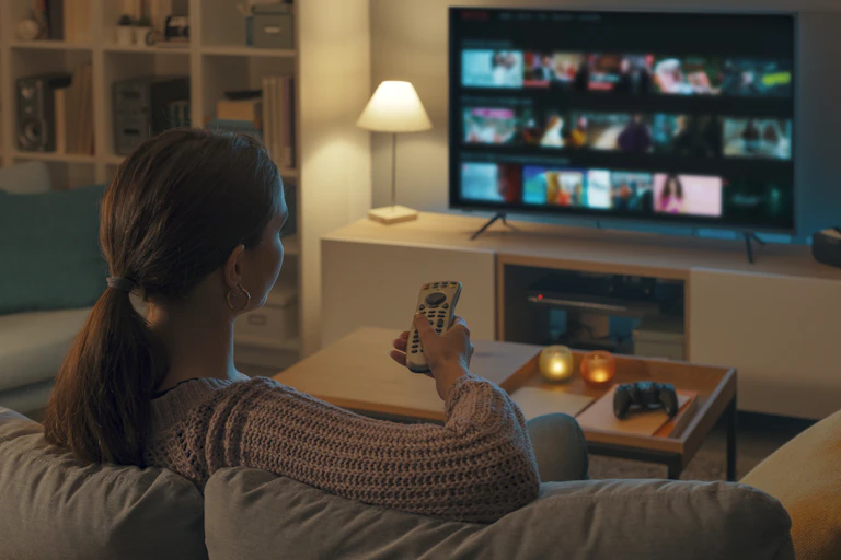 Woman relaxing on the couch, she is using the remote control and choosing a TV show or movie on the television menu
