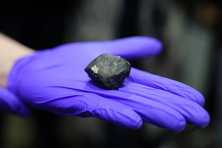 A gloved hand holding what looks like a small rock that is black on the outside with a grey interior