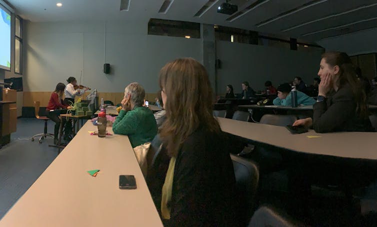 Students seen sitting in a university classroom as a person plays the violin.