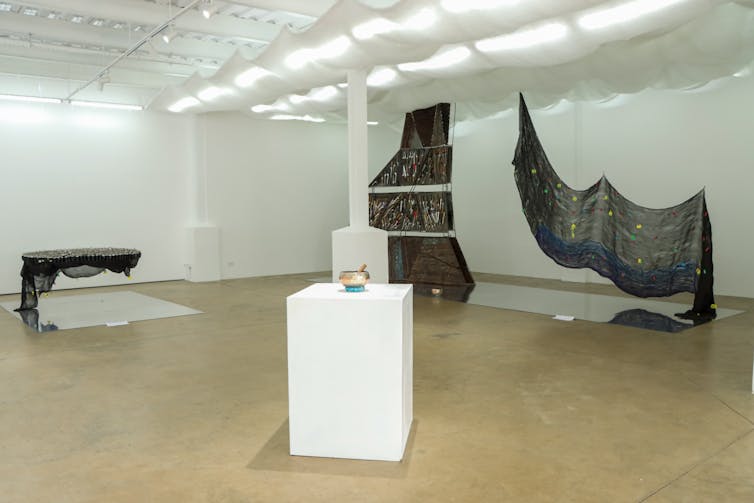 A view of an art exhibition, a pedestal in the foreground holds a shiny gold bowl. In the background cloys and metal sculptures hang above mirrors.