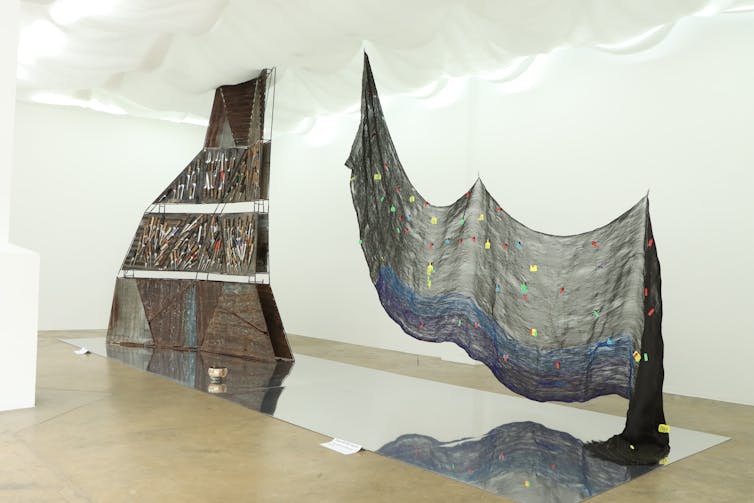 Two large sculptures are installed above mirrors, one a cloth with coloured objects attached, the other a rusted metal sheet with knives attached.