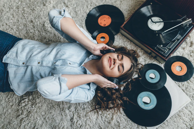 A young woman, lying down on a carpet, with vinyl records around her, smiles as she listens to music through her headphones.