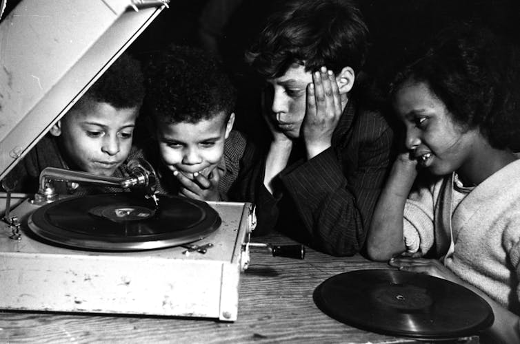 A young Black woman and three children smile as they listen to music on a record player.
