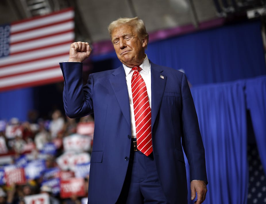 A middle-aged white man dressed in a dark business suit raises his fist as he walks on stage underneath a large American flag.