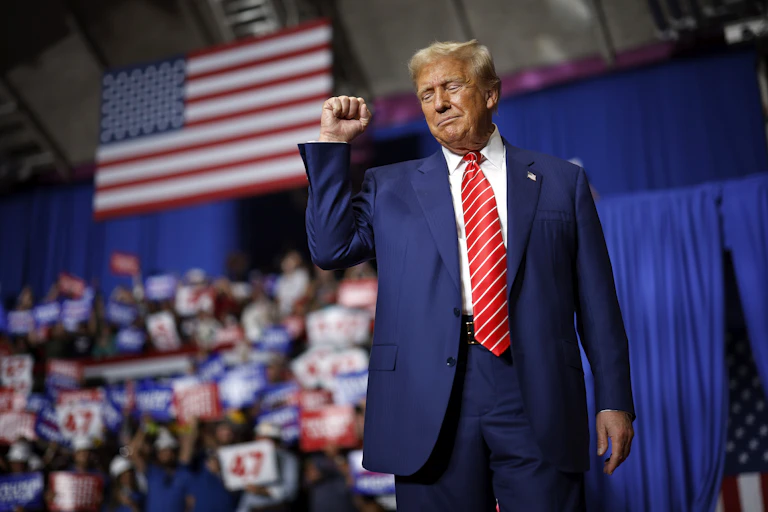 A middle-aged white man dressed in a dark business suit raises his fist as he walks on stage underneath a large American flag.