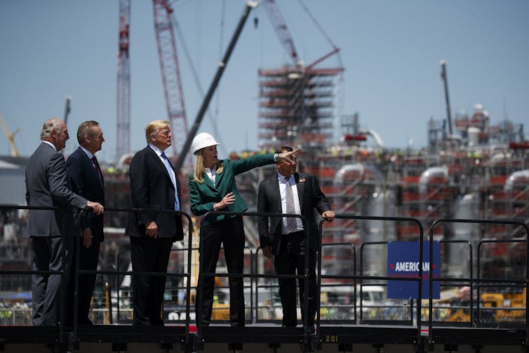 Trump stands with men in suits and a woman wearing a hard hat cranes behind them.