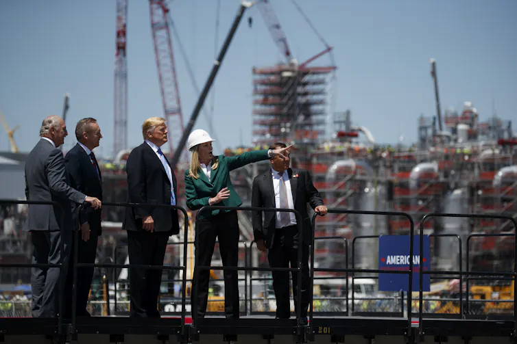 Trump stands with men in suits and a woman wearing a hard hat cranes behind them.