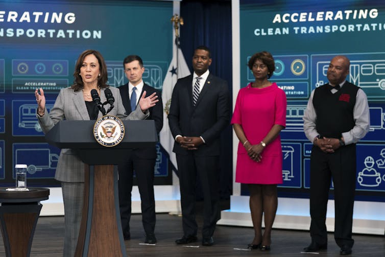 Harris stands with members of her cabinet and a bus driver in front of a sign discussing clean transportation.