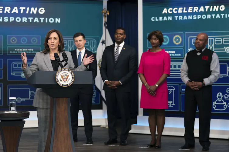Harris stands with members of her cabinet and a bus driver in front of a sign discussing clean transportation.