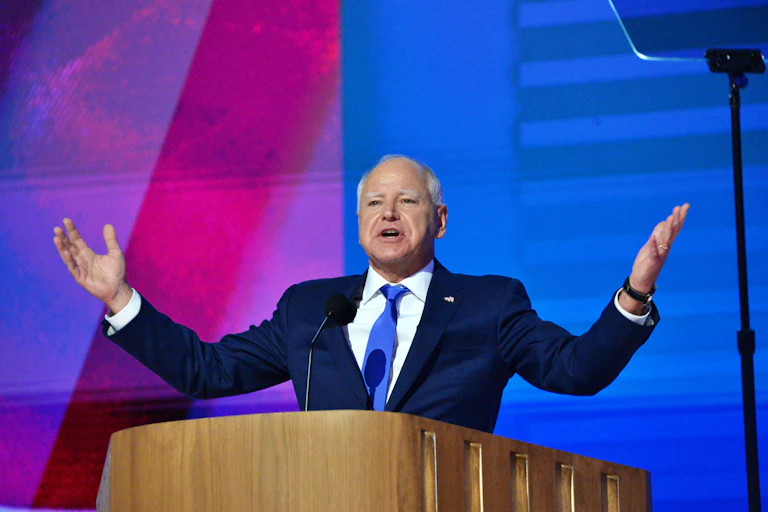 A man stands at a lectern on a stage with his arms extended.