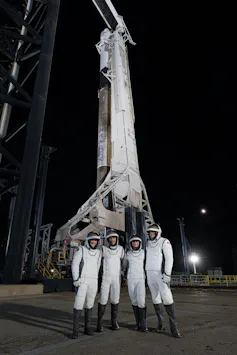 Empat astronaut mengenakan setelan putih dan helm berdiri di depan roket di landasan peluncuran.