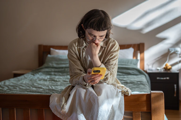 Young woman sitting on a bed, holding a phone and looking scared.