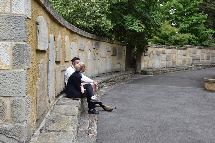 Two people sit alongside a memorial wall in a park