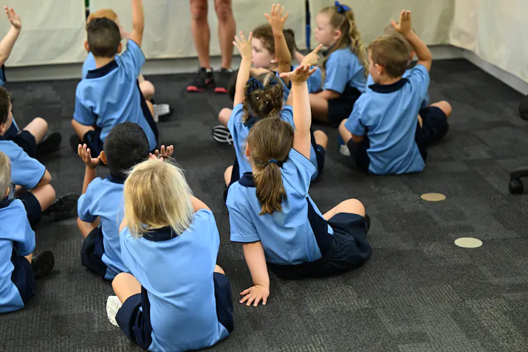 Young children in uniform sit on the floor and raise their hands.