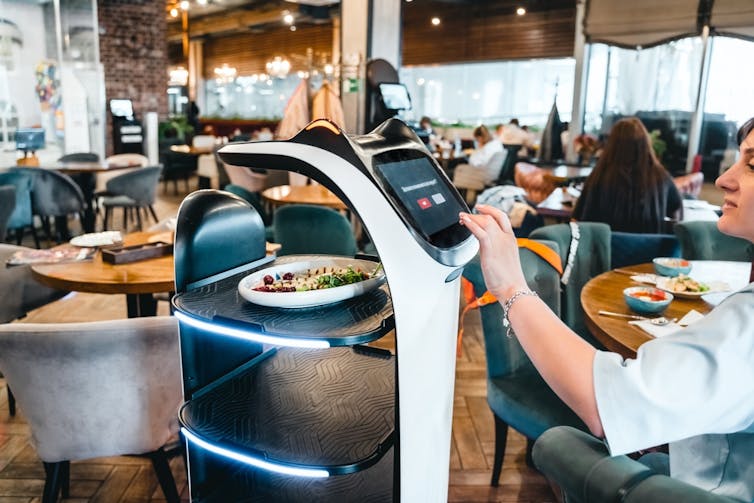 A person at a restaurant interacts with a delivery robot that has shelves for food and cute cat ears above a screen.