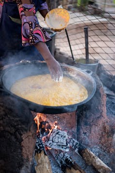 A woman's arm can be seen cooking a palm oil bean cake over a fire in Nigeria