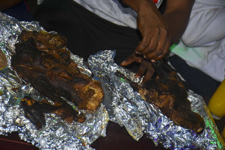 Two hands opening a tin foil package of cooked goat meat that has been cooked on a fire