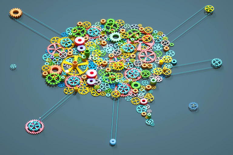 A digitally generated image of multi coloured gear wheels connected together in the shape of a brain on grey background