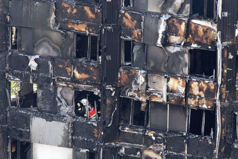 The burnt outside of a building with a firefighter visible through one of the windows.