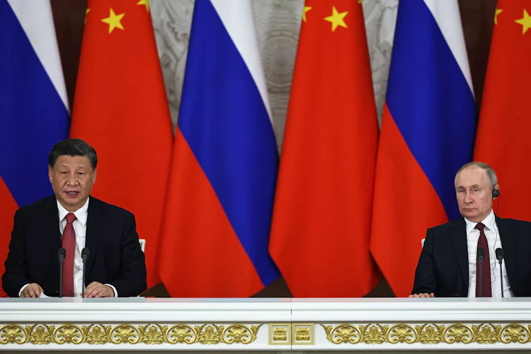Two men wearing suits and ties address the media while sitting, withj their backs to RUussian and Chienes flags.