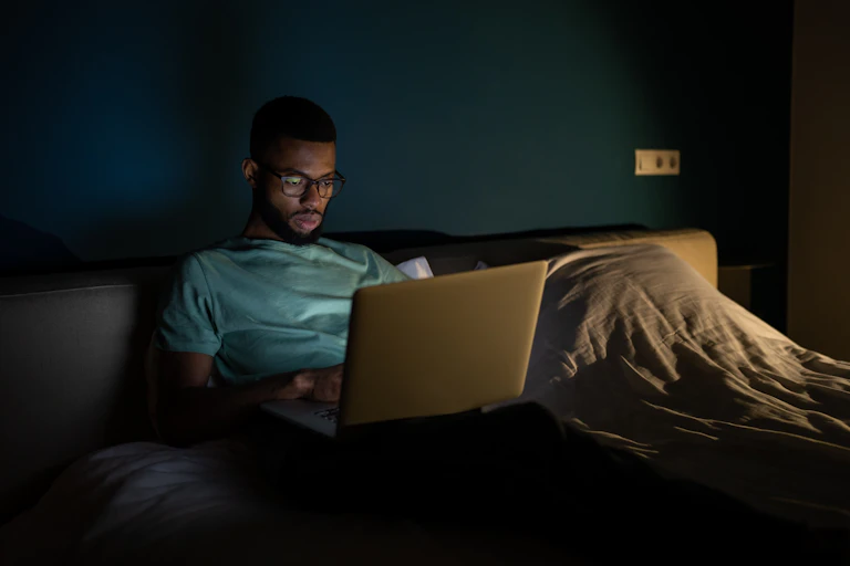 man in bed in dark room looking at laptop