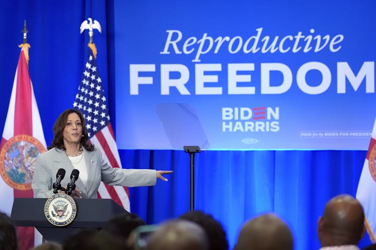 A dark-haired woman in a grey suit jacket stands on a stage behind a lectern with a banner behind her that reads Reproductive Freedom.