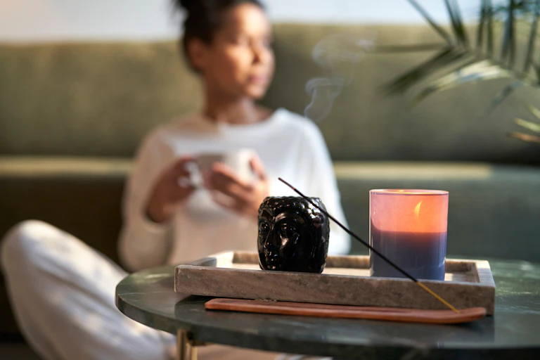 Une femme floue en arrière plan avec une tasse à la main. Au premier plan, une bougie d'ambiance et de l'encens brûlent.