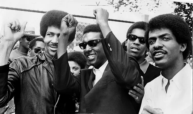 Several young Black men, wearing dark glasses, smile as they raise their fists.