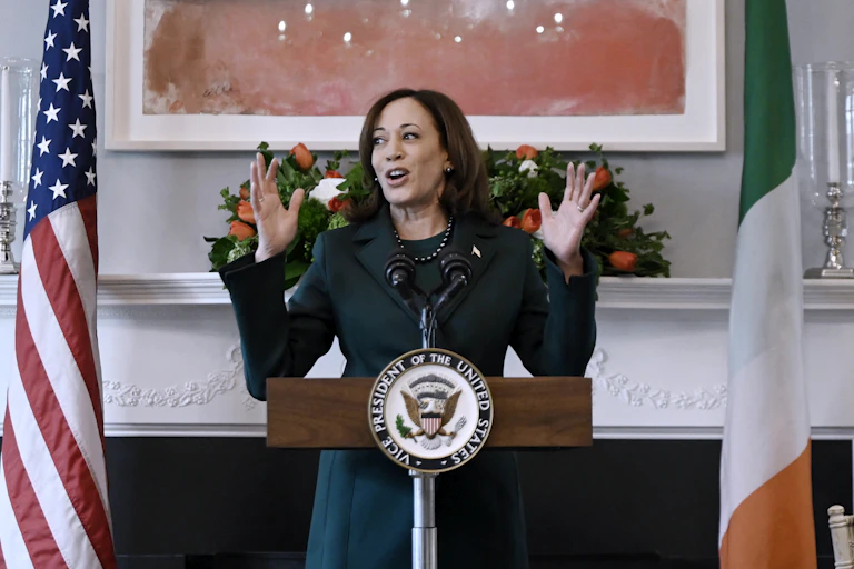 A woman in a green suit stands at a lectern flanked by an American and Irish flag.